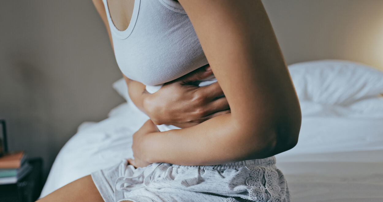 Vrouw die op bed zit met haar armen om haar buik. Ze heeft buikpijn door stress.