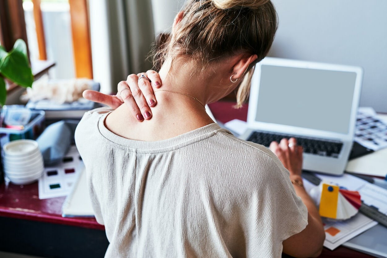 Vrouw die aan het werk is achter haar laptop. Ze masseert haar nek, omdat ze last heeft van nekpijn.