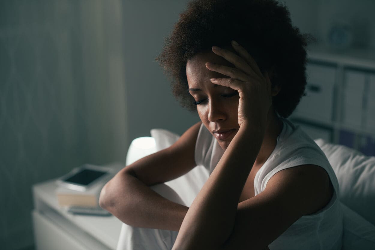 Vrouw die in het donker in haar slaapkamer op bed zit. Z heeft last van spanningshoofdpijn.