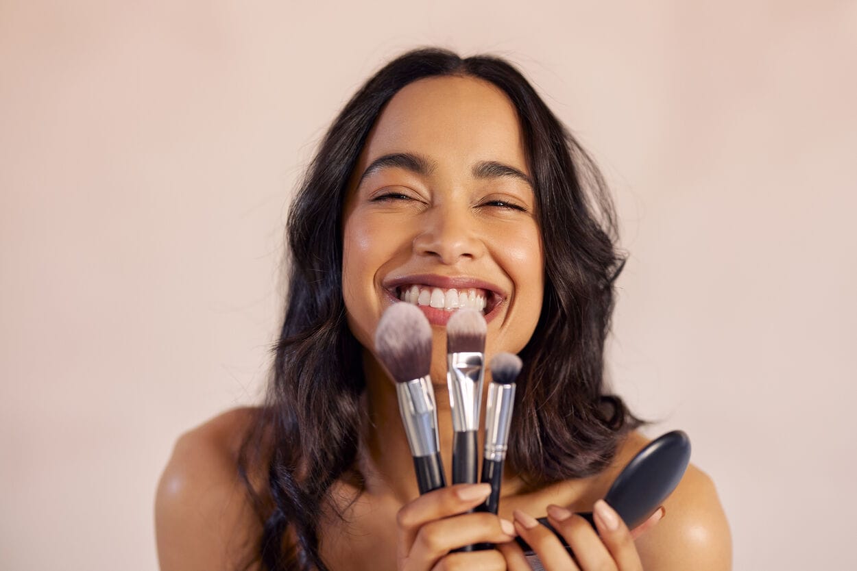 Vrouw met een roze achtergrond die meerdere make-upkwasten in haar hand heeft. Ze gaat de juiste volgorde van make-up toepassen.
