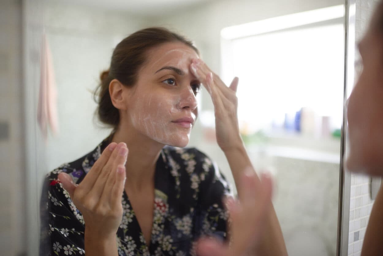 Vrouw voor de spiegel in de badkamer. Ze smeert een cleanser op haar gezicht.