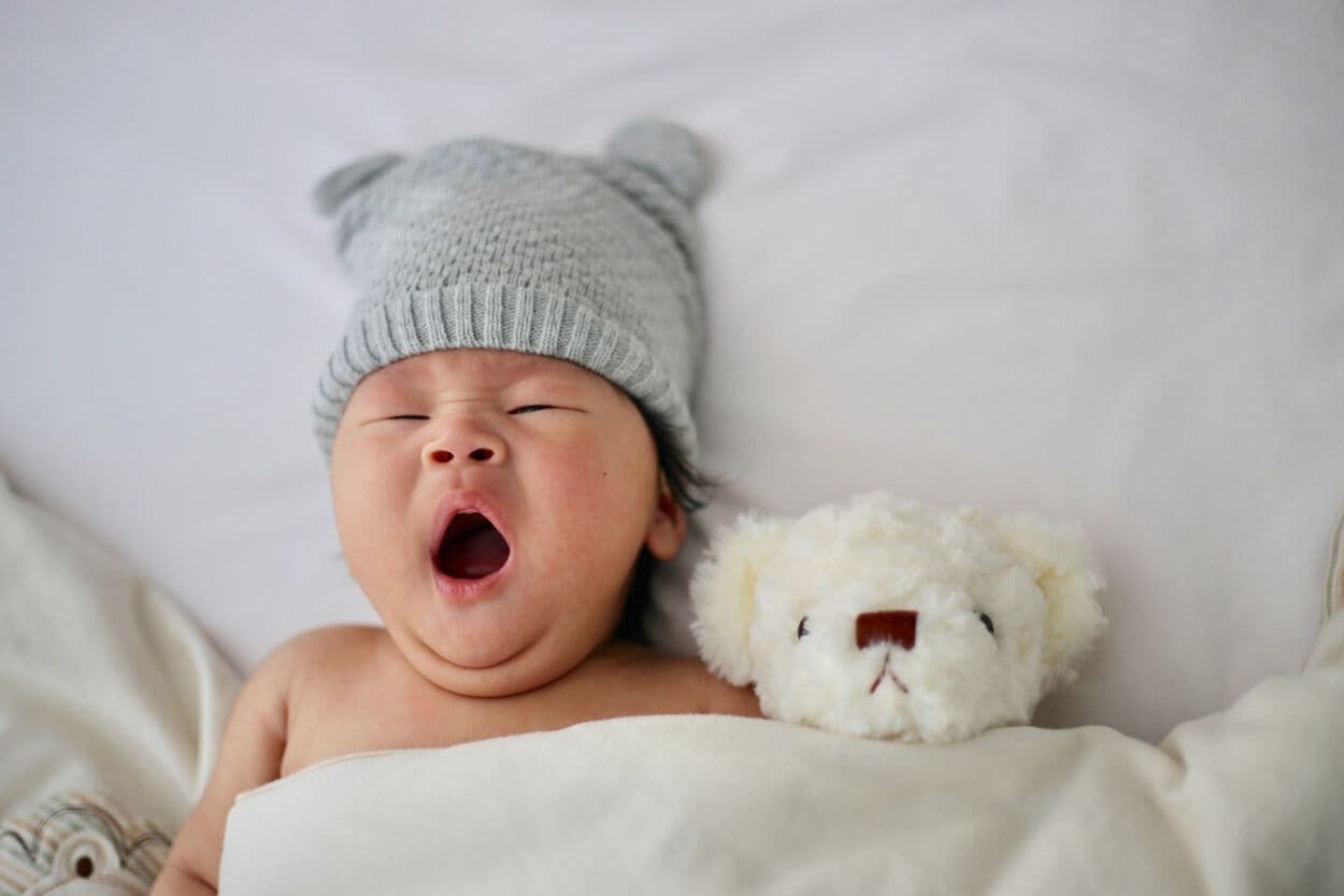 Jonge baby inbed onder dekentje met muts op en beertje naast zich